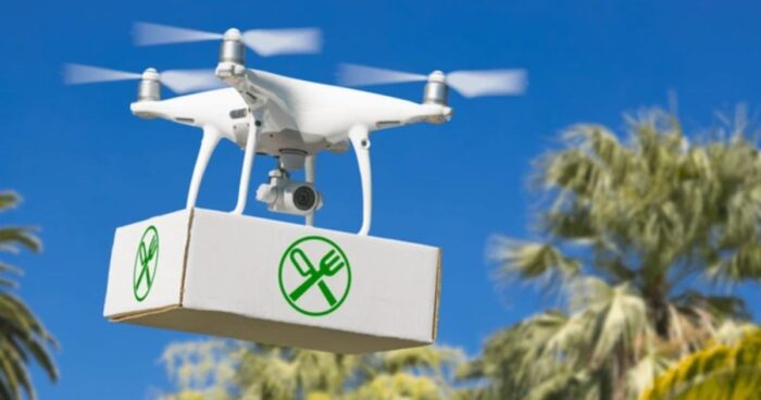 futuro-food-delivery-drone-pizza-robot Un drone consegna una pizza a un robot su un balcone di un palazzo cittadino, rappresentando il futuro del food delivery